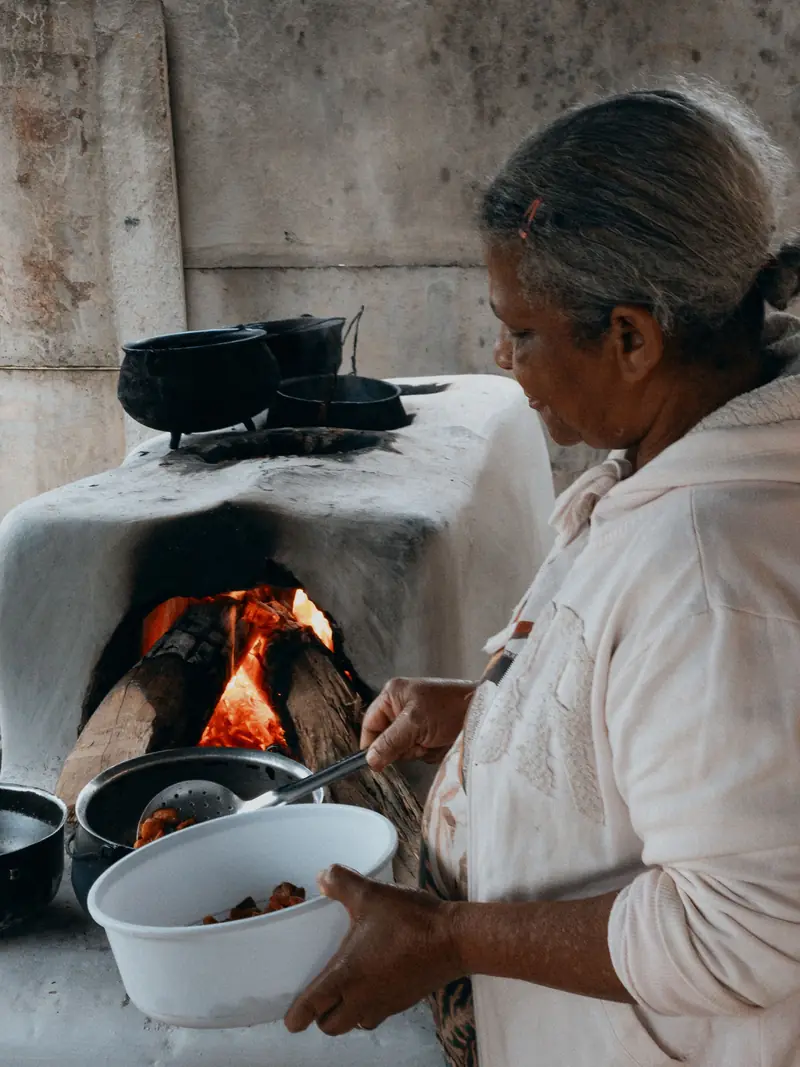 Foto ilustrativa: mulher serve alimento de uma panela em um fogão a lenha, em uma cozinha tradicional