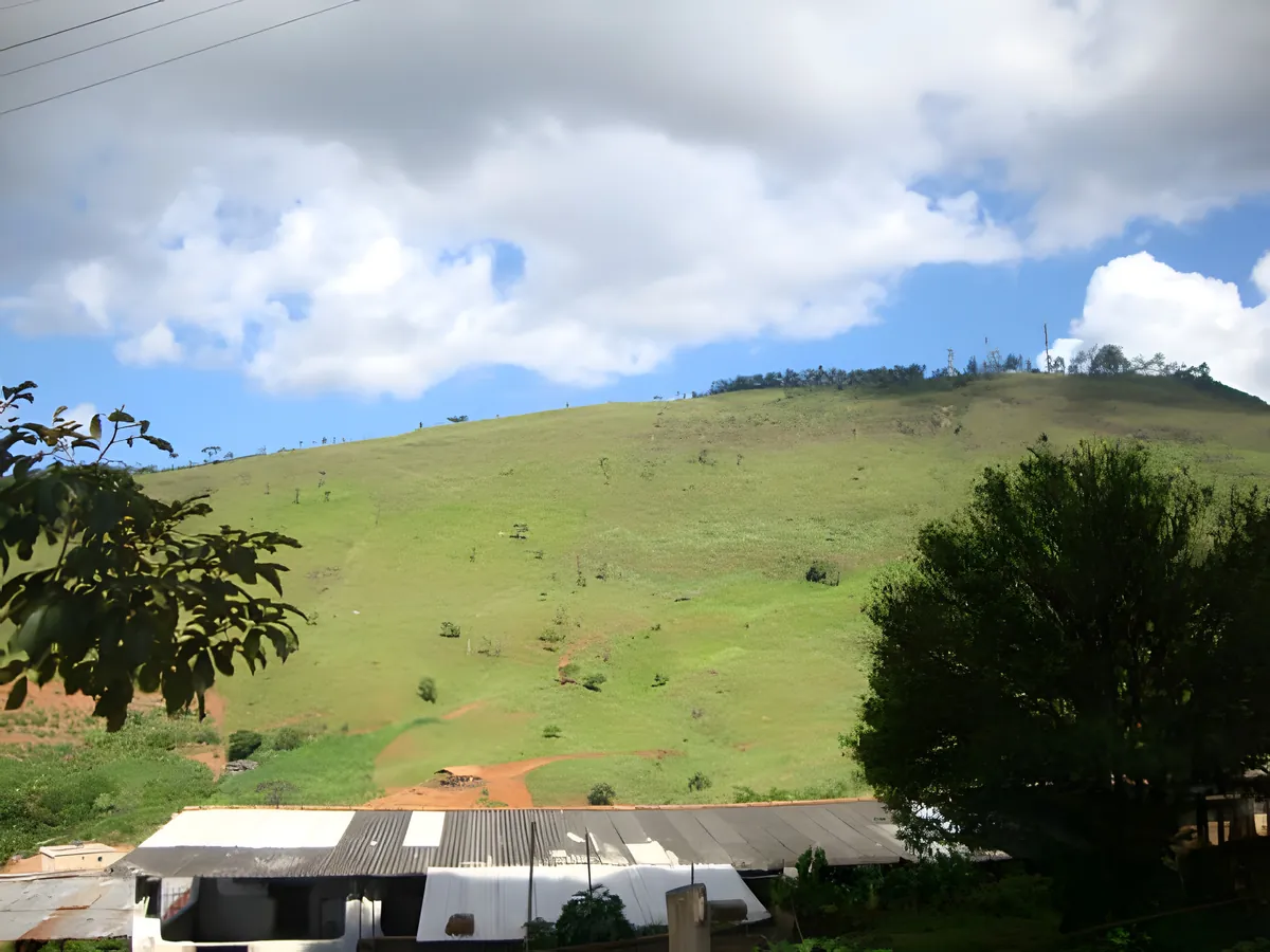 Foto real: colina verde sob céu azul com nuvens, vista da Colônia, com telhados no pé do morro