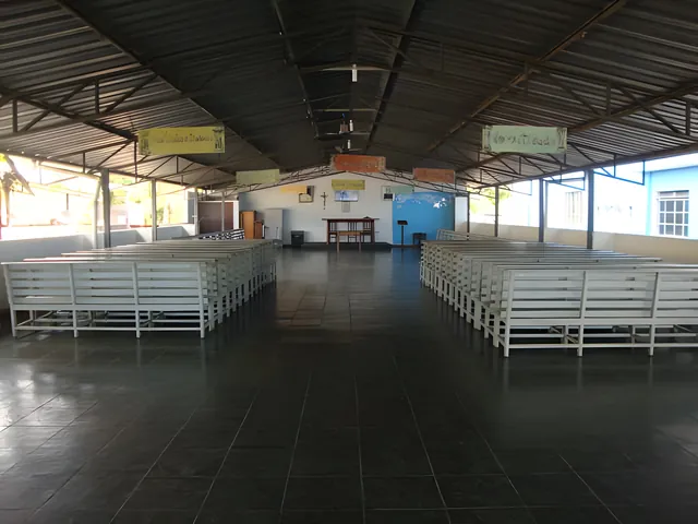 Foto real: salão coberto da Colônia, com bancos brancos em fileiras e, ao fundo, uma mesa de altar com crucifixo