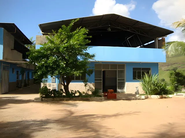 Foto real: prédio azul de dois níveis da Colônia Bom Samaritano, com uma árvore e o pátio de terra